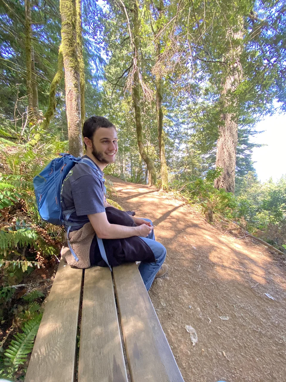 Ty smiling on a bench along a wooded coastal trail